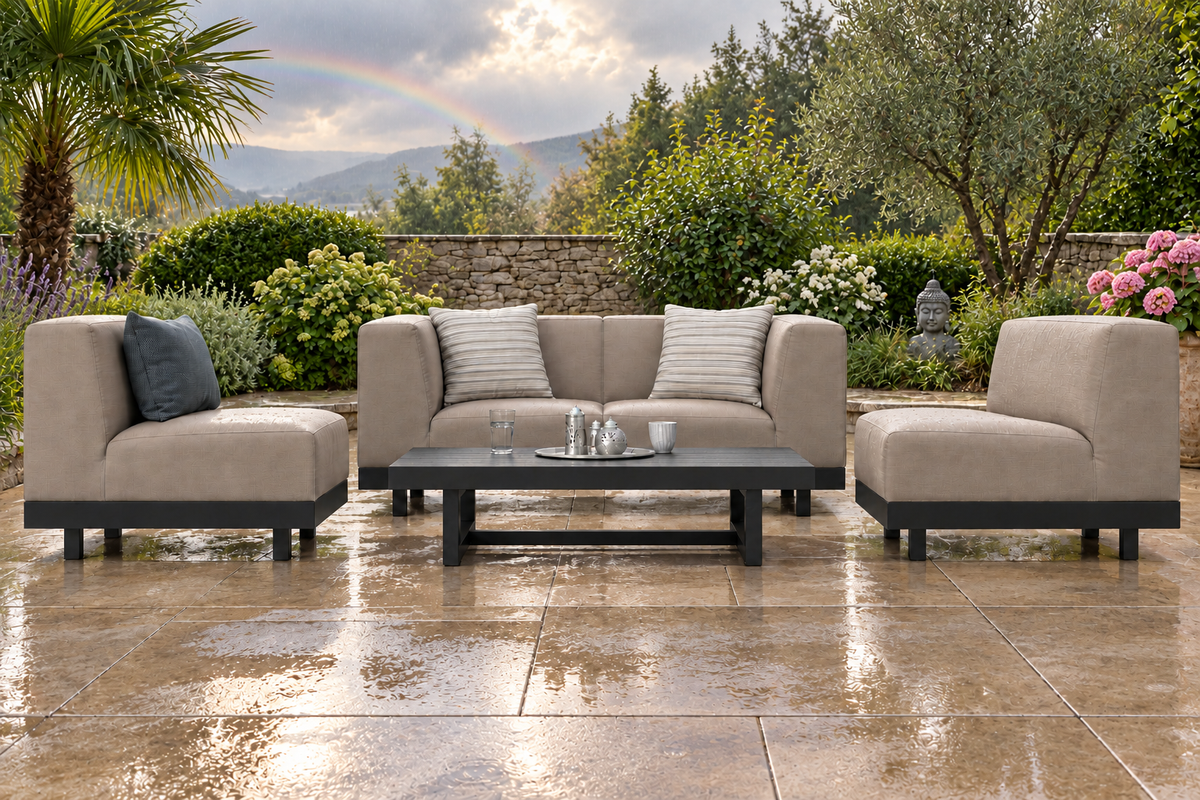 Die Silan Gartenlounge steht auf einer breiten, regennassen Terrasse, umgeben von viel Grün und einigen rosa Blüten. Im Hintergrund erkennt man einen ganz zarten Regenbogen über sanften Hügeln.